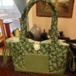 A green floral-patterned fabric bag with two handles and a decorative button sits on a wooden surface in front of a framed picture.