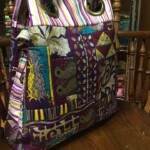 Colorful patchwork tote bag with a mix of patterns and stripes, featuring large eyelets for handles, placed on a wooden chair.