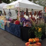Market stall with colorful handmade bags and textiles displayed on tables under a white tent. Pumpkins and flowers decorate the stand.