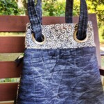 A fabric handbag with spiral patterns on top and solid blue bottom rests on a wooden bench outdoors.