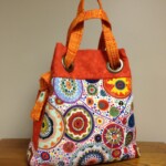 Colorful handbag with circular patterns and orange handles, resting on a wooden table against a plain background.