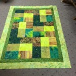 A handmade quilt with green, blue, and brown patchwork squares on a carpeted floor.