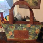 Fabric tote bag with floral and brown patterns, placed on a wooden surface near spools of thread and a framed tree picture.