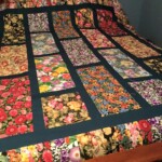 A bed covered with a colorful patchwork quilt featuring various floral patterns, surrounded by a dark blue border.