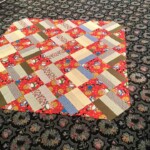 A patchwork quilt with colorful patterns is laid on a patterned carpeted floor.