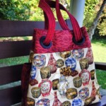 A fabric bag with red handles and a pattern featuring various colorful vases sits on a wooden bench outdoors.
