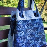 Blue and white patterned bag with large handles resting on a wooden bench outdoors, surrounded by greenery.