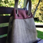 A gray herringbone-patterned tote bag with brown accents rests on a wooden bench outside.