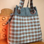Plaid tote bag with brown, blue, and white checkered pattern, featuring ring handles and a bow on the side, on a wooden surface.