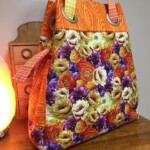A colorful tote bag with large floral patterns and orange side panels, placed on a wooden table next to a small cabinet.