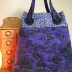 Purple floral-patterned tote bag with round handles, placed next to a small wooden drawer on a wooden surface.