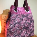 A pink and purple tote bag with a leaf and dot pattern, featuring large grommets and dark handles, placed on a wooden surface.
