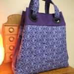 Purple and white patterned tote bag with large handles, resting on a wooden surface near a small chest of drawers.