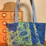 Colorful paisley-patterned handbag with long handles, displayed in front of a small wooden drawer unit.