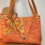 An orange and yellow patterned handbag with a large V-flap and button, featuring braided straps, displayed on a wooden surface.
