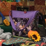 A colorful fabric bag with a butterfly design sits among various textiles, sunflowers, and a small orange pumpkin.