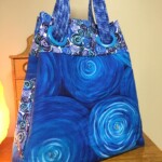A blue tote bag with swirling circular patterns sits on a wooden table near a wooden chest.