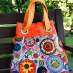 A colorful tote bag features vibrant circular patterns and orange handles, resting on a wooden bench in a lush garden setting.