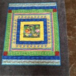 Colorful quilt with floral patterns and animal motifs, bordered by geometric designs, placed on a gray textured surface.