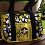 A handbag with bee-themed patterns and a gold bee emblem on the front, resting on a wooden bench.