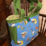 Colorful fabric tote bag with green patterned handles and blue cartoon-themed body, placed on a wooden chair.