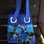 A blue and purple tote bag with floral patterns and fabric handles, placed on a brown wooden bench.