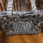 A fabric bag with blue and gray feather patterns on a wooden floor, featuring two handles and an exterior pocket.