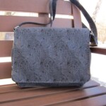 A dark patterned handbag is placed on a wooden bench under sunlight.