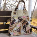 A floral-patterned handbag sits on a wooden bench outdoors, surrounded by autumn leaves.
