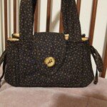 A small, dark patterned fabric handbag with a button closure and short handles, resting on a pink surface.