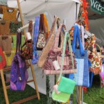 A display of colorful handmade bags hanging on a wooden ladder at an outdoor market stall, with jars on a table nearby.