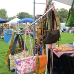 A display of colorful handmade bags hanging at an outdoor craft fair, with tents and people in the background.