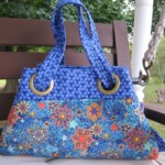A blue handbag with brass ring handles and a colorful star pattern rests on a wooden swing outdoors.