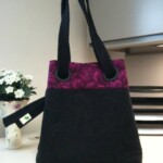 A black tote bag with a purple floral top edge and two handles is displayed on a table near a small flowerpot.