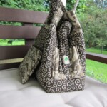 A patterned bag with swirls and a pocket sits on an outdoor bench with greenery in the background.