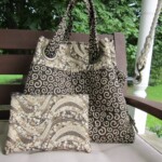 A matching patterned handbag and clutch with swirls and floral designs on a bench, set against a backdrop of green foliage.