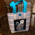 Striped tote bag with a colorful cat design and blue handles, placed on a wooden chair.
