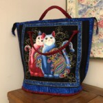 A colorful tote bag with an embroidered design of two ornate cats, one red and one blue, resting on a wooden surface.