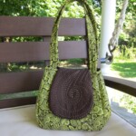 A green and brown patterned handbag sits on a wooden bench with a garden in the background.