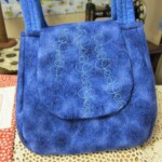 A blue fabric handbag with a circular stitched pattern is displayed on a textured surface.