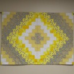 A quilt with a geometric pattern in yellow, white, and blue tones hangs on a wall.