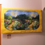 Colorful landscape painting with a garden path, framed in yellow, hanging on a wall near a doorway.