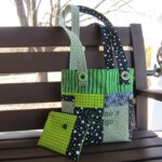 A patchwork tote bag and matching wallet with green, black, and white patterns on a wooden bench.