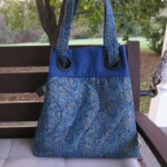 A blue and patterned tote bag rests on a wooden bench outdoors, with trees and green grass in the background.
