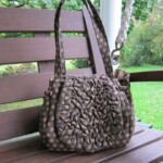 A brown polka dot handbag with ruched fabric detail, resting on a wooden bench with greenery in the background.