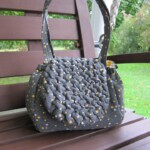 A textured, dark gray handbag with yellow dots is placed on a wooden bench outdoors.