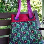 A colorful handbag with purple handles, adorned with a floral pattern, rests on a wooden bench in a garden setting.