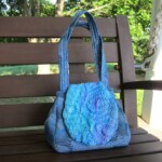 A blue knitted handbag with a textured circular flap rests on a wooden bench outdoors.