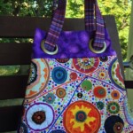 A colorful tote bag with circular patterns and purple handles, resting on a wooden bench outdoors.