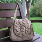 A textured beige handbag with floral patterns is displayed on a wooden bench outdoors.